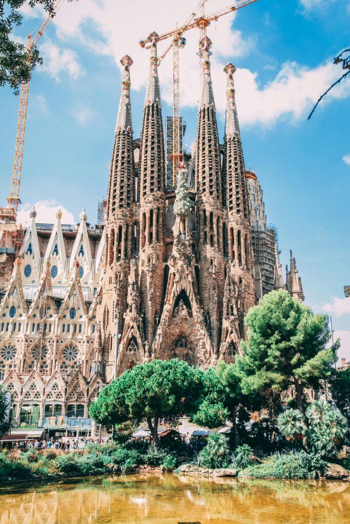 External shot of the visually stunning Sagrada Familia in Barcelona. The fantastically ornate cathedral dominates the picture. In the foreground is a pond and mature trees.